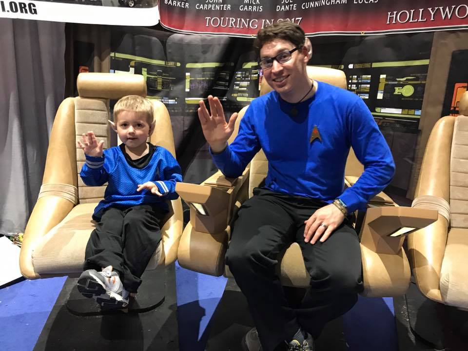 Dad and son in matching Star Trek Spock cosplay, flashing the Live Long and Prosper hand signal while sitting on the bridge of the Enterprise