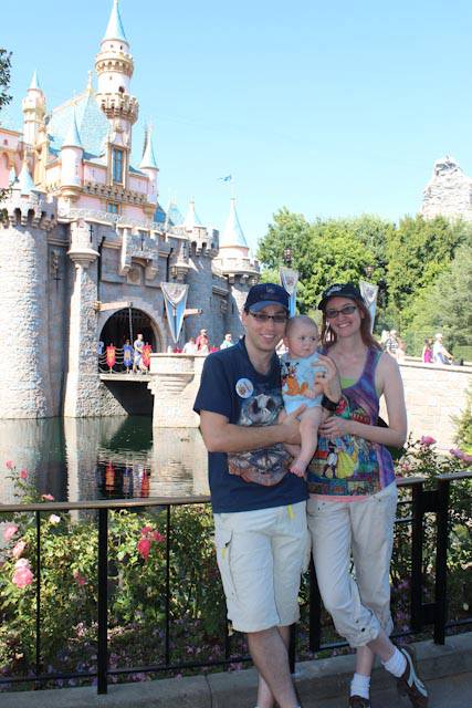 A family visiting Disneyland with their baby, standing in front of Sleeping Beauty's castle