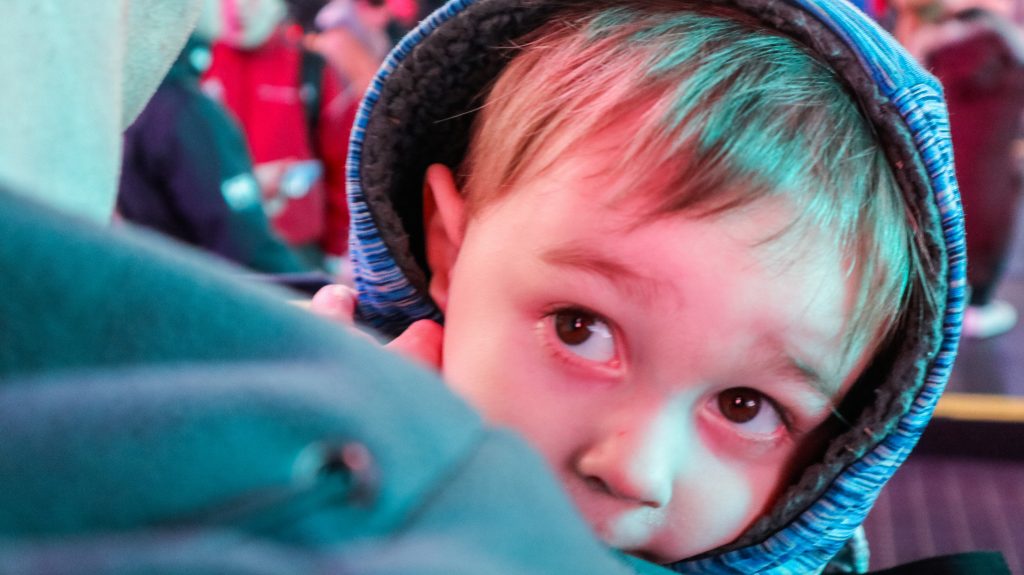 A toddler boy peeking his head over dad's shoulder, wearing a coat hood