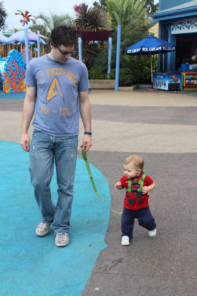 A dad and his toddler son, wearing a green backpack with a leash