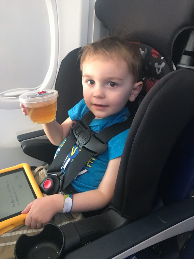 A little boy in a car seat on an airplane holds a cup of juice and a tablet on his lap