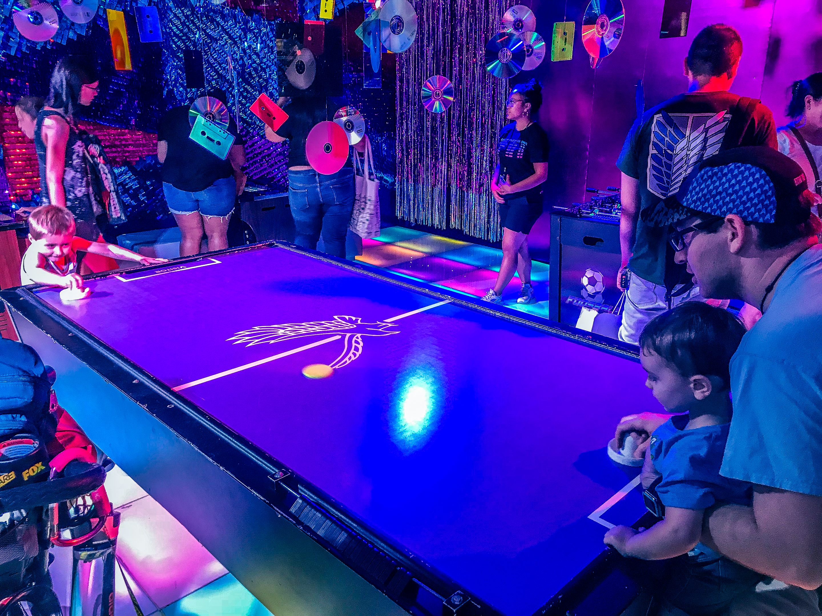 A family plays air hockey in a purple lit room, themed like Ready Player One at SDCC